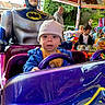 adult, amusement_park, background, batman, blue_jacket, child, colorful, fun, hat, lights, outdoor, park, people, playful, ride, smile, statue, steering_wheel, toddler, toy_car