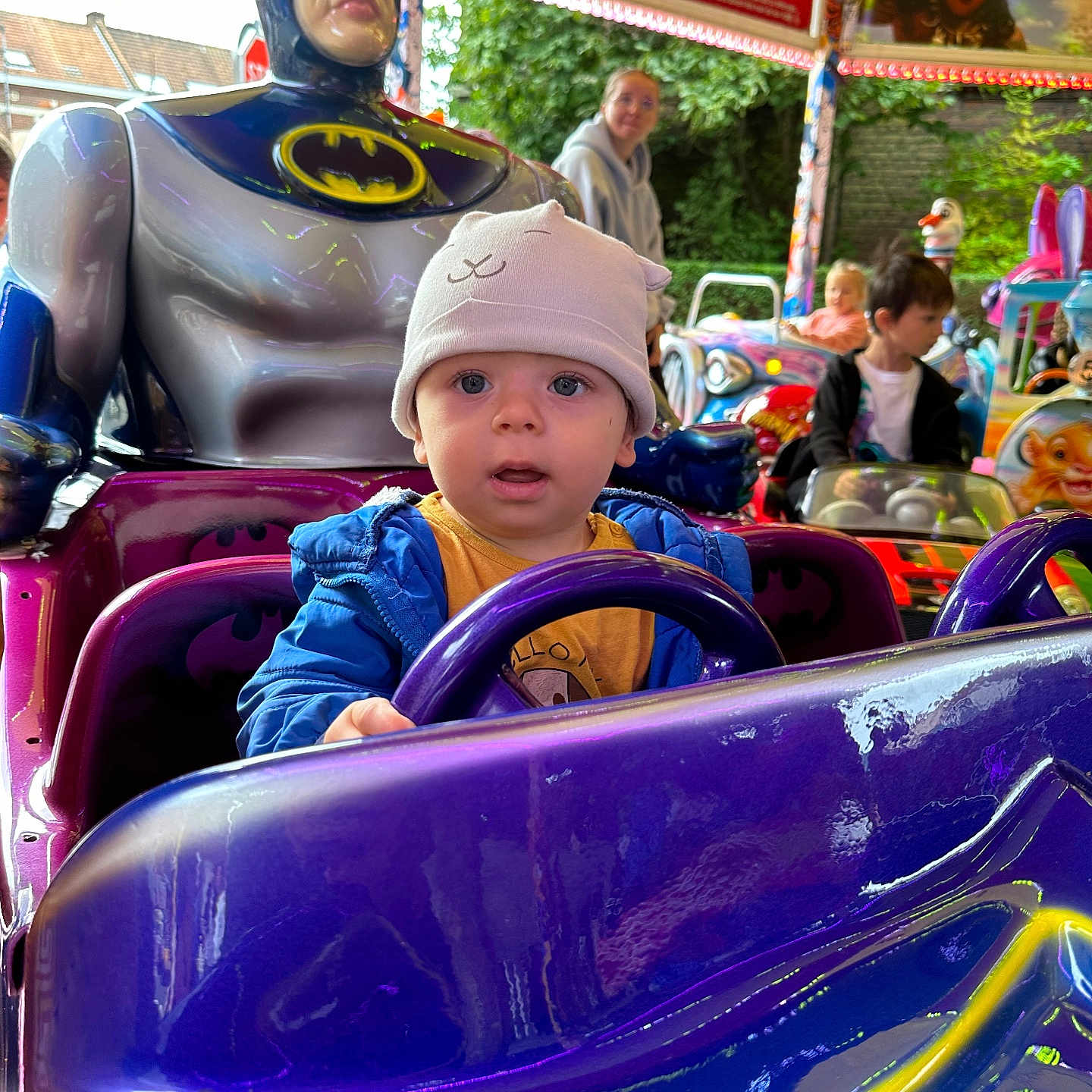 Isaac participe au concours pour gagner de l'argent avec cette photo : toddler, child, batman, ride, amusement_park, toy_car, hat, blue_jacket, steering_wheel, statue, adult, smile, outdoor, colorful, lights, fun, playful, background, people, park