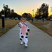 Devina joined the competition — help win amazing prizes! toddler, child, cow_onesie, walking, sidewalk, sunset, park, trees, grass, street_lights, cars, outdoor, cute, person, footwear, casual_clothing, expression, young_child, daylight, nature