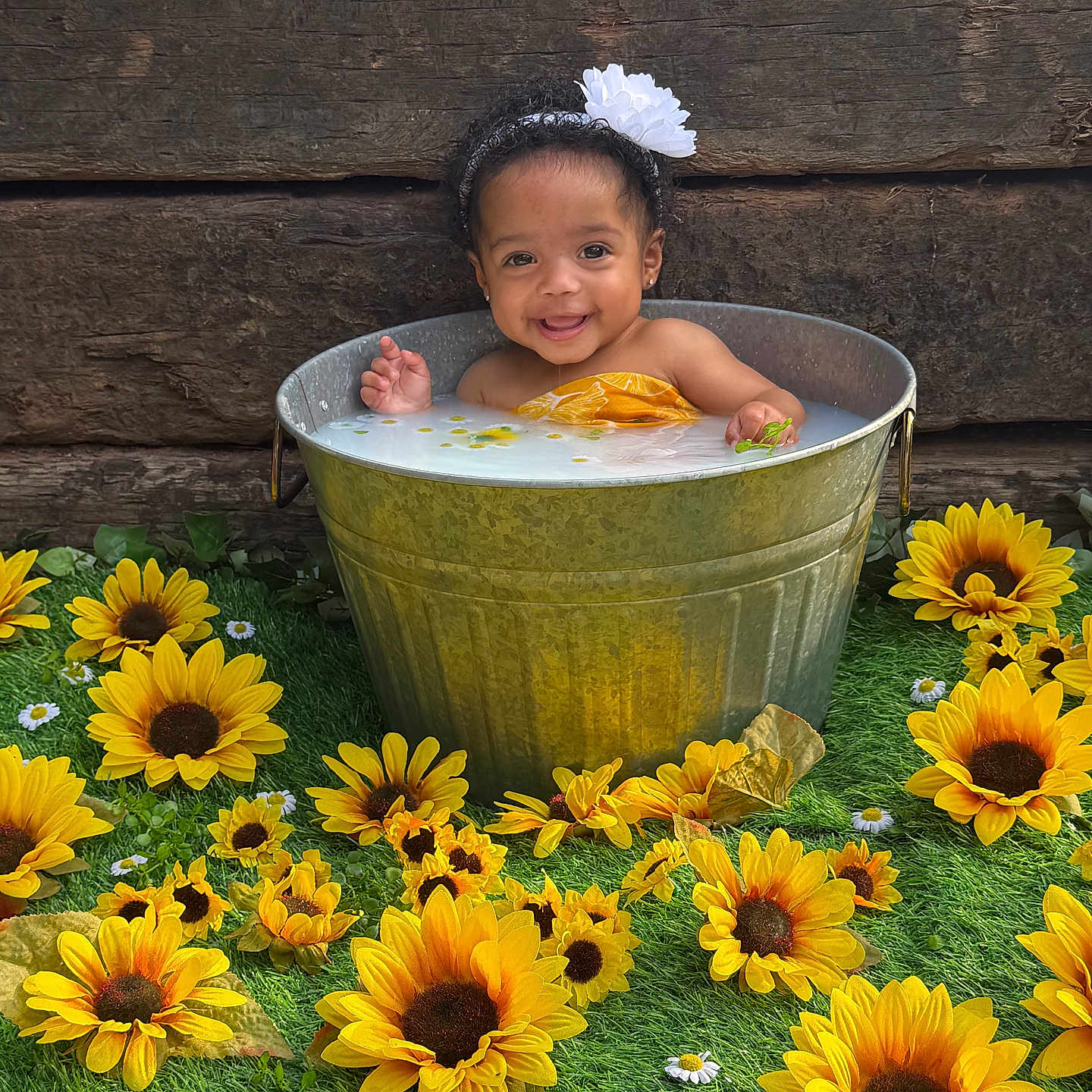 Alea is registered to the contest to win money with this photo: baby, bucket, child, cute, flower, flower_arrangement, grass, greenery, happy, headband, infant, milky_water, nature, outdoor, portrait, smiling, sunflower, water, wood, yellow