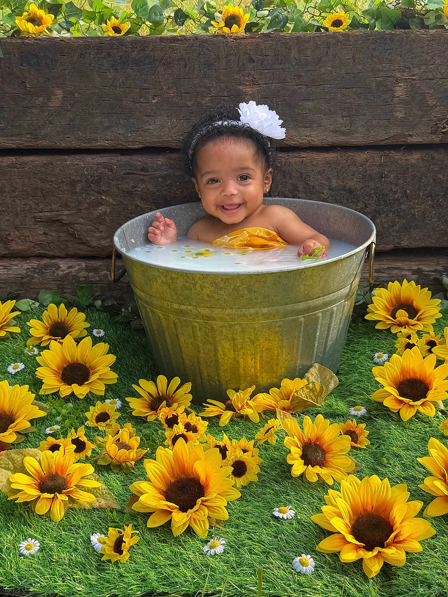 Alea is registered to the contest to win money with this photo: baby, child, bucket, flower, sunflower, grass, wood, headband, smiling, water, milky_water, greenery, nature, portrait, outdoor, cute, happy, infant, yellow, flower_arrangement
