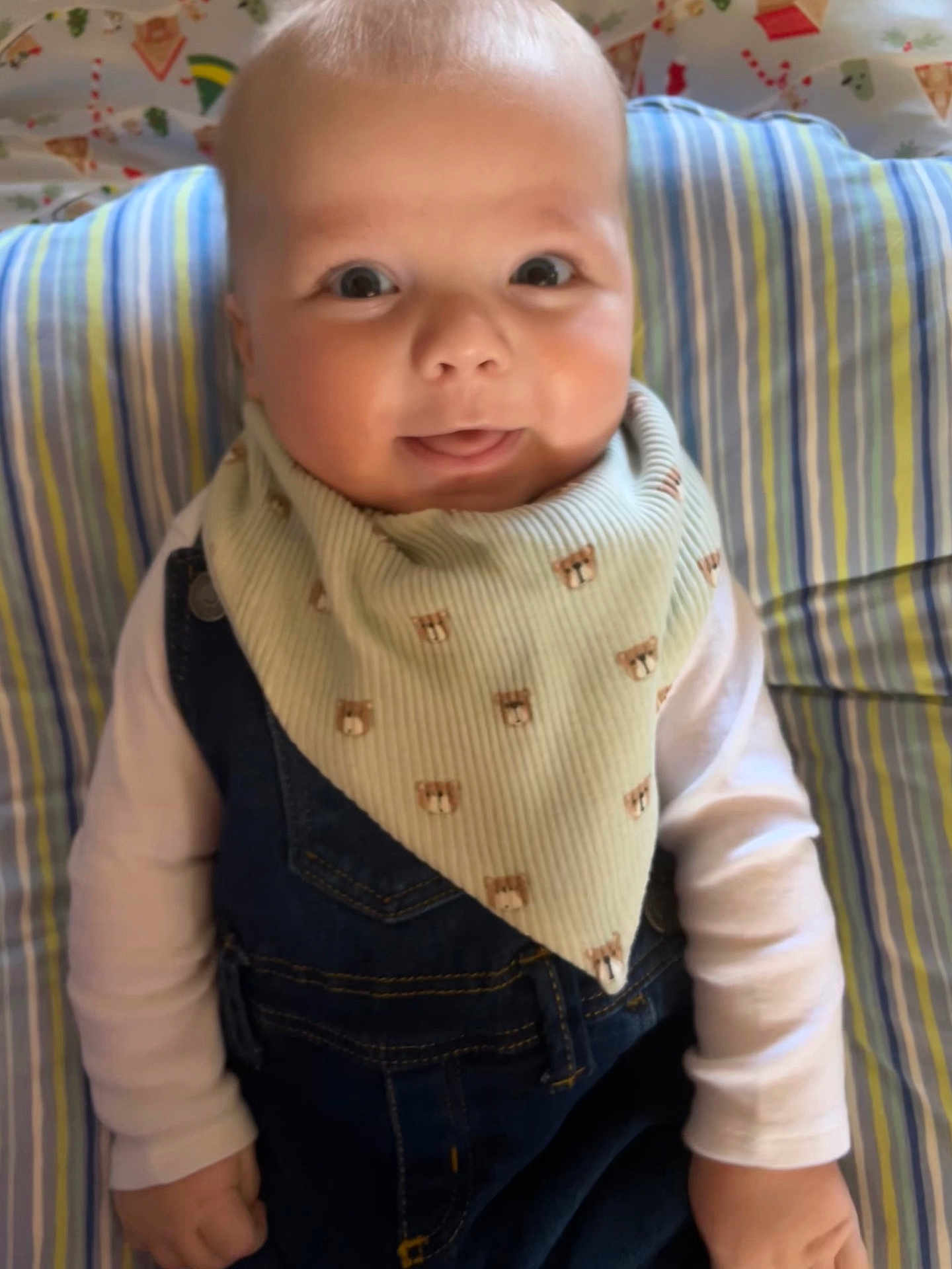 Makai is registered to the contest to win money with this photo: baby, child, infant, denim_overalls, bib, striped_cushion, smiling, tongue_out, cute, indoors, portrait, happy, young_child, clothing, person, seated, background_pattern, face, head, hands
