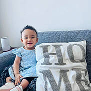 Georgina is registered to the contest to win money with this photo: baby, blanket, blue_dress, casual, child, comfortable, couch, cozy, curly_hair, furniture, happy, home_decor, indoor, person, pillow, portrait, shoes, sitting, smiling, toddler