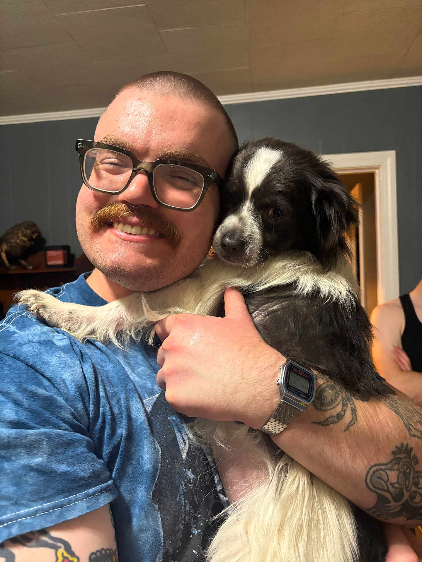 Big Ed joined the competition — help win amazing prizes! man, dog, glasses, mustache, tattoo, watch, smile, hug, indoor, blue_shirt, black_and_white_dog, short_hair, person, background, doorway, casual, happy, pet, friendship, home