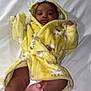 baby, child, yellow_robe, bed, white_bedspread, foot, face, curly_hair, infant, cute, lying_down, cozy, blanket, diaper, sleepwear, indoors, soft_fabric, smiling, skin, hooded_clothing