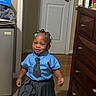 child, toddler, girl, school_uniform, striped_shirt, tie, pleated_skirt, sneakers, white_socks, hair_beads, blue_accessories, standing, smiling, indoors, bedroom, door, dresser, carpet, mini_fridge, portrait