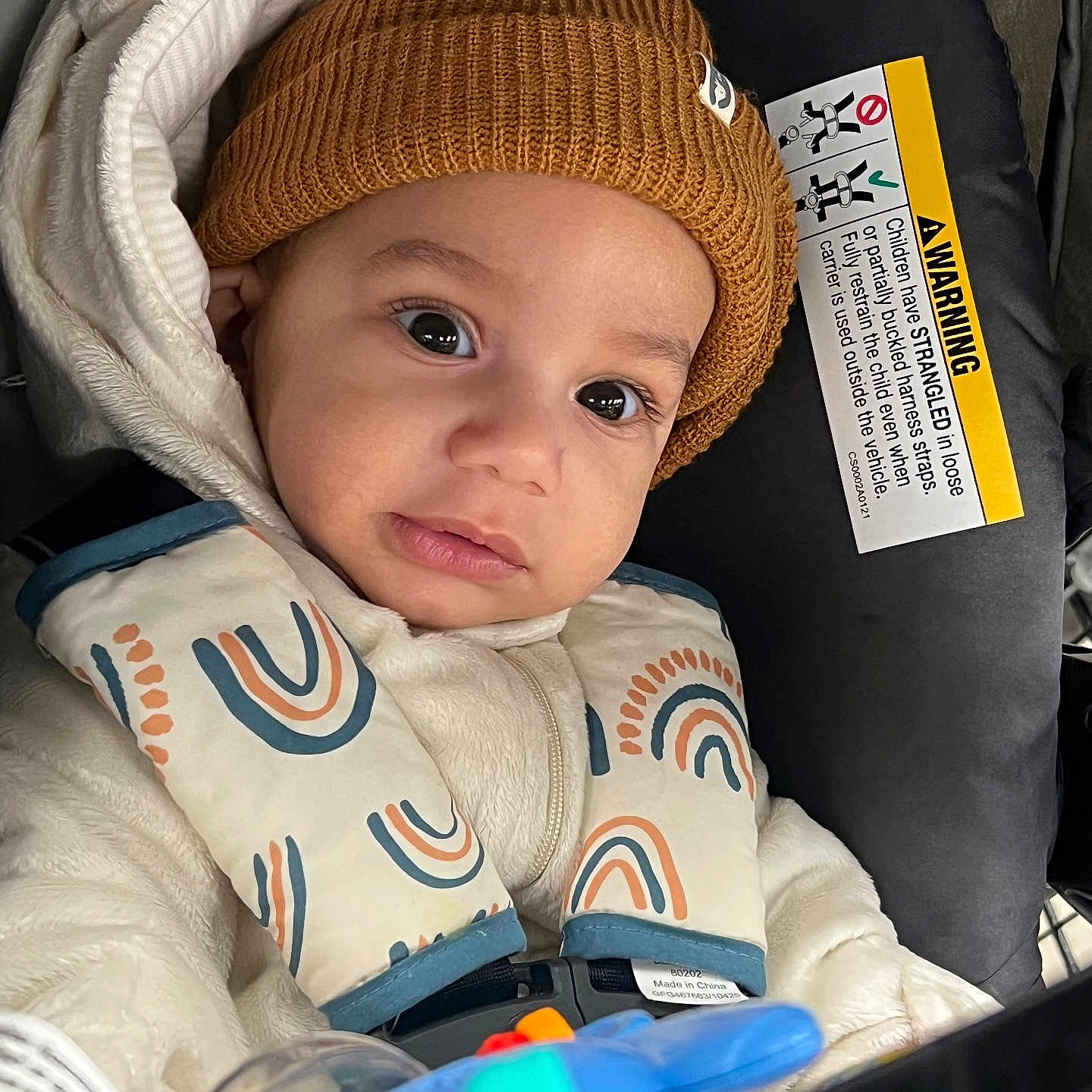 Zyon joined the competition — help win amazing prizes! baby, child, car_seat, hat, knit_hat, jacket, rainbow_pattern, toy, blue_toy, infant, closeup, face, eyes, cute, warm_clothing, seatbelt, car, portrait, indoor, child_safety
