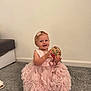 child, baby, girl, pink_dress, flower_clips, carpet, indoor, smiling, holding_object, footwear, sitting, cute, decorative_sign, happy, portrait, head, person, floor, wall, home