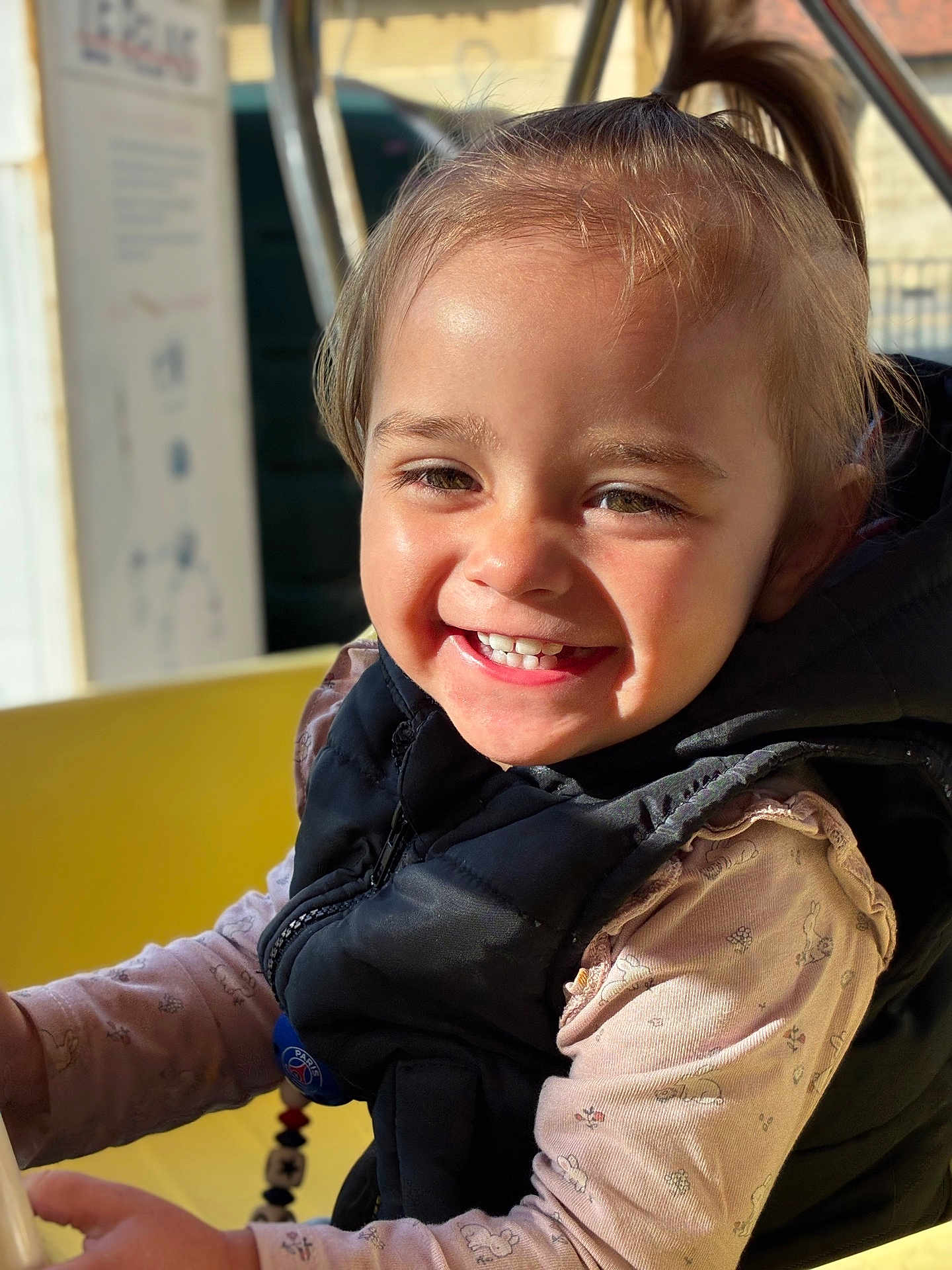 Lyana participe au concours pour gagner de l'argent avec cette photo : toddler, child, smiling, face, vest, long_sleeve_shirt, outdoor, sunlight, happy, person, portrait, cute, young_child, holding, playground, blurry_background, daylight, clothing, hair, skin
