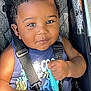 baby, child, smiling, stroller, seatbelt, toy, curly_hair, blue_shirt, red_shorts, closeup, infant, cute, happy, sitting, outdoor, sunlight, black_hair, person, portrait, baby_toy