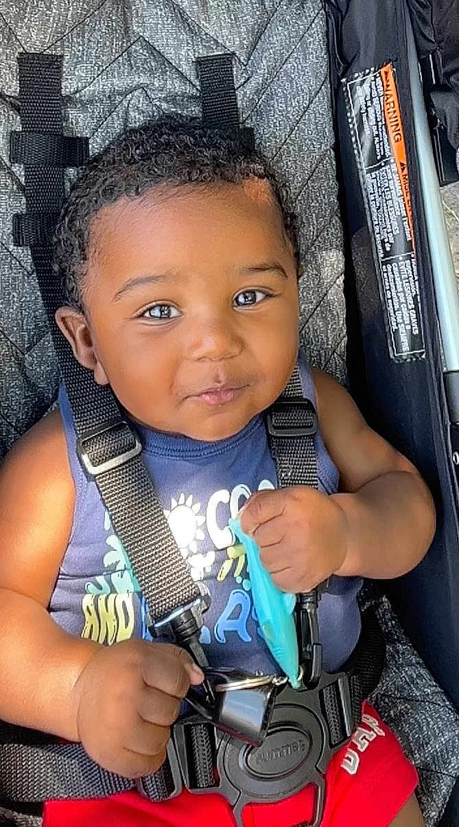Yani joined the competition — help win amazing prizes! baby, child, smiling, stroller, seatbelt, toy, curly_hair, blue_shirt, red_shorts, closeup, infant, cute, happy, sitting, outdoor, sunlight, black_hair, person, portrait, baby_toy