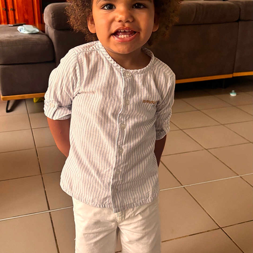 Soën a rejoint le concours — aidez-le/la à gagner de superbes lots ! cartoon, child, crocs, curly_hair, cute, footwear, furniture, happy, indoor, living_room, person, smiling, sofa, standing, striped_shirt, television, tile_floor, toddler, white_shorts, young_child