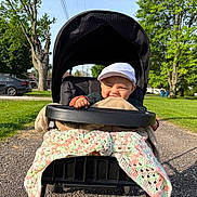 Elijah is registered to the contest to win money with this photo: baby, stroller, blanket, outdoor, greenery, trees, sunlight, smile, child, park, grass, pathway, cap, happy, person, nature, daytime, car, residential, relaxation