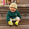 baby, child, smiling, bench, hat, green_jacket, socks, wooden_bench, urban, brick_wall, shop_window, happy, cute, portrait, seated, outdoor, infant, casual, colorful, playful