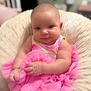 Fernanda is registered to the contest to win money with this photo: baby, infant, child, pink_dress, frills, cushion, soft_texture, indoor, portrait, cute, chubby, smiling, sitting, person, closeup, skin, face, hand, comfort, cozy