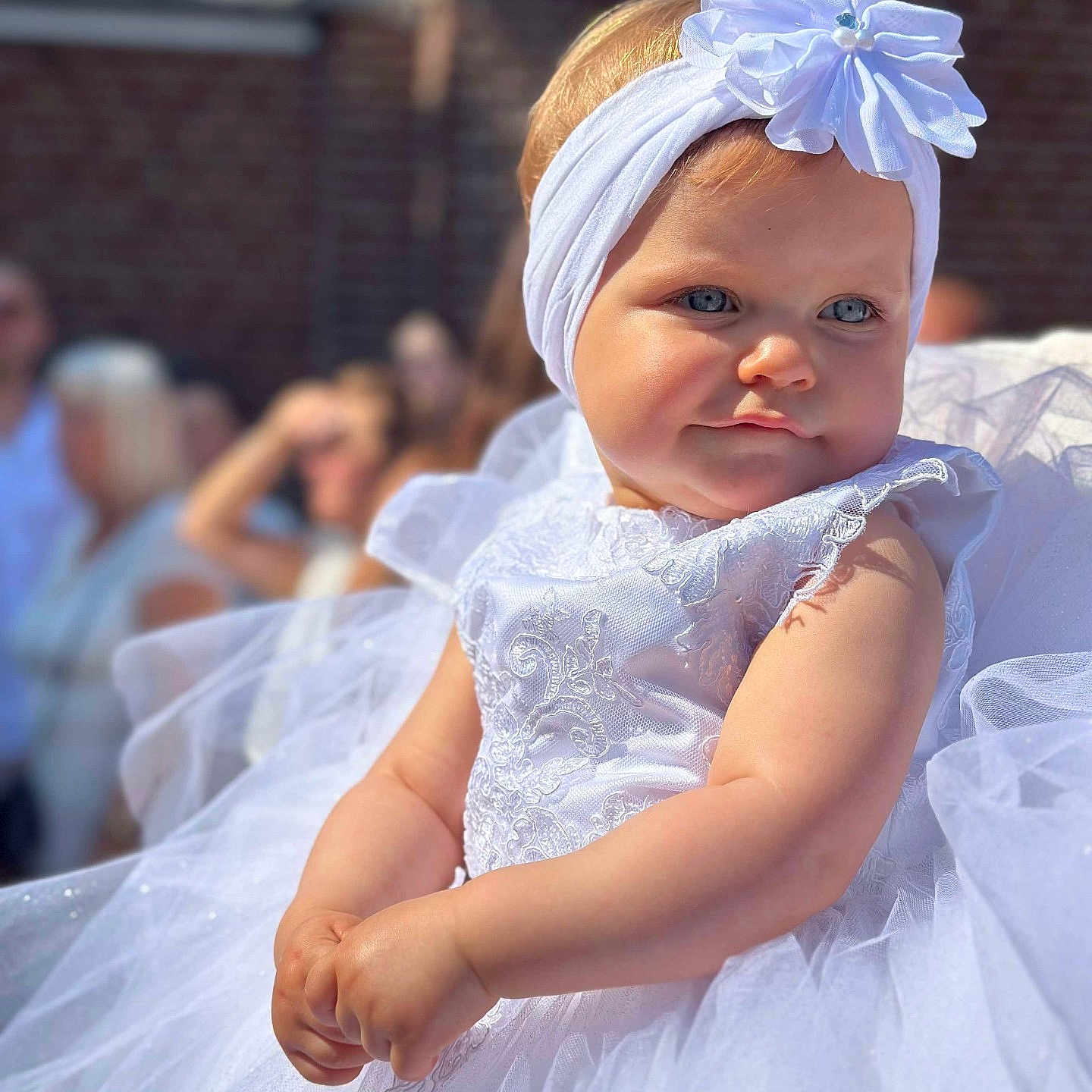 Aislyne a rejoint le concours — aidez-le/la à gagner de superbes lots ! baby, blue_eyes, celebration, child, cute, dress, event, face, flower, hands, headband, infant, outdoor, people_in_background, portrait, smile, soft_focus, sunlight, tulle, white_dress