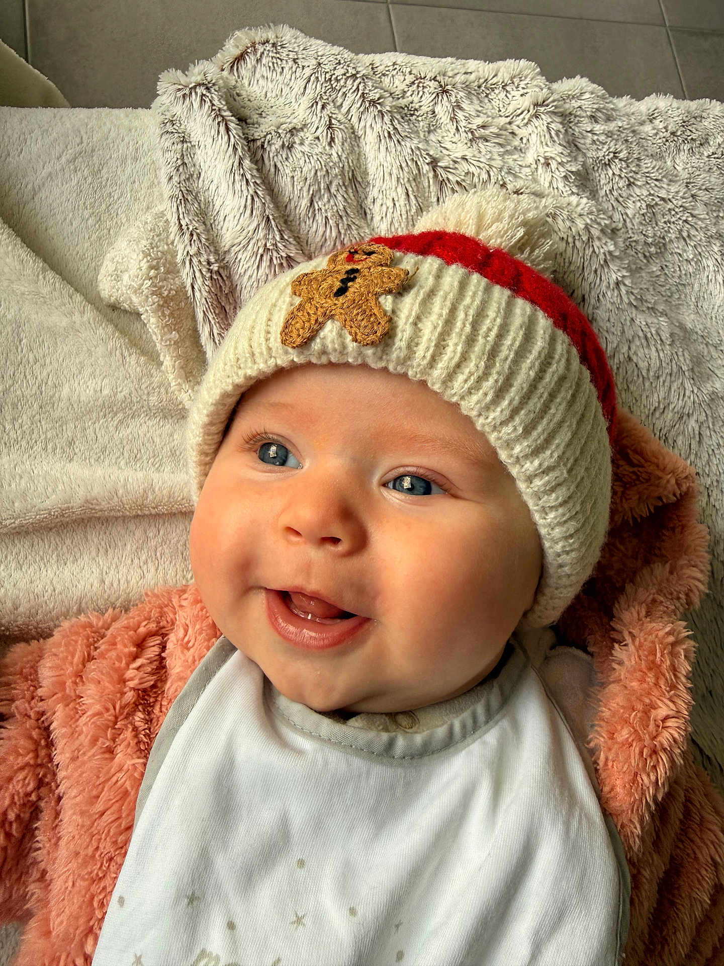 Olivia participe au concours pour gagner de l'argent avec cette photo : baby, infant, knitted_hat, gingerbread_patch, smile, tongue_out, blue_eyes, cheeks, bib, blanket, plush_texture, fur_blanket, cozy, portrait, close_up, laying_down, happy, cute, warm_clothing, indoor