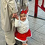 toddler, child, adult, holding_hands, red_bow, red_skirt, white_jacket, scarf, sidewalk, outdoor, footwear, happy, walking, person, pavement, winter_clothing, smiling, urban, family, support