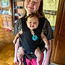 baby, child, baby_carrier, smiling, indoor, person, face, happy, pacifier, black_shirt, pink_pants, home_interior, wooden_floor, wooden_furniture, decor, portrait, holding, parenting, family, cute