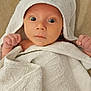 baby, towel, hooded_towel, white_towel, infant, cute, wide_eyes, skin, face, hands, raised_fists, soft_texture, beige_background, wrapped, newborn, portrait, indoor, child, expression, blanket