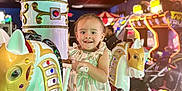 Mila is registered to the contest to win money with this photo: toddler, child, carousel, arcade, horse, ride, striped_dress, smiling, indoor, colorful_lights, amusement, playful, toy, entertainment, game_machines, person, fun, happy, decorative, family