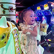 Mila is registered to the contest to win money with this photo: toddler, child, carousel, horse, arcade, neon_lights, indoor, ride, fun, smiling, pigtails, dress, toy, amusement, game, colorful, playful, happy, entertainment, person