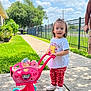 Mila is registered to the contest to win money with this photo: toddler, child, toy_stroller, plush_toys, sidewalk, grass, tree, fence, blue_sky, clouds, white_shoes, red_pants, winnie_the_pooh, shirt, outdoor, sunny, person, smiling, daytime, play