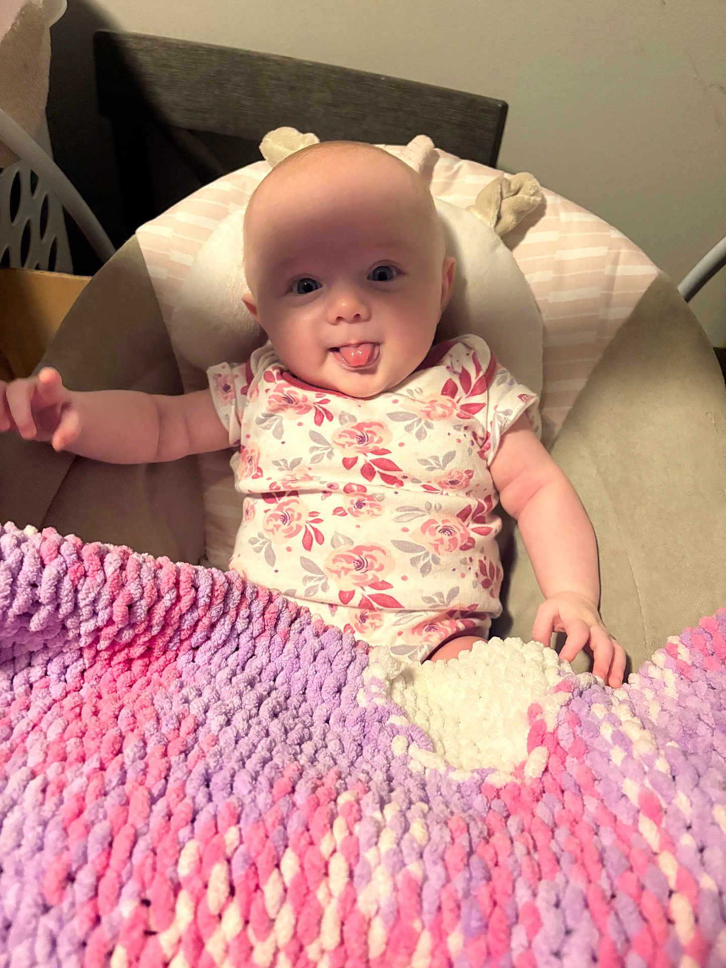Adelina is registered to the contest to win money with this photo: baby, infant, tongue_out, blanket, knitted_blanket, floral_onesie, cushion, chair, indoor, cute, playful, child, person, portrait, smiling, soft_texture, home, cozy, seated, happy