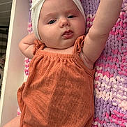Adelina is registered to the contest to win money with this photo: baby, infant, headband, orange_romper, knitted_blanket, purple, pink, cute, chubby_cheeks, lying_down, reaching_arm, indoors, soft_texture, toddler, child, person, portrait, closeup, expression, headwear