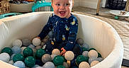 Weston is registered to the contest to win money with this photo: baby, child, ball_pit, toys, indoor, smiling, red_hair, onesie, playtime, colorful_balls, infant, floor, furniture, sitting, happy, person, home, activity_center, basket, shelf