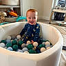 baby, child, ball_pit, toys, indoor, smiling, red_hair, onesie, playtime, colorful_balls, infant, floor, furniture, sitting, happy, person, home, activity_center, basket, shelf