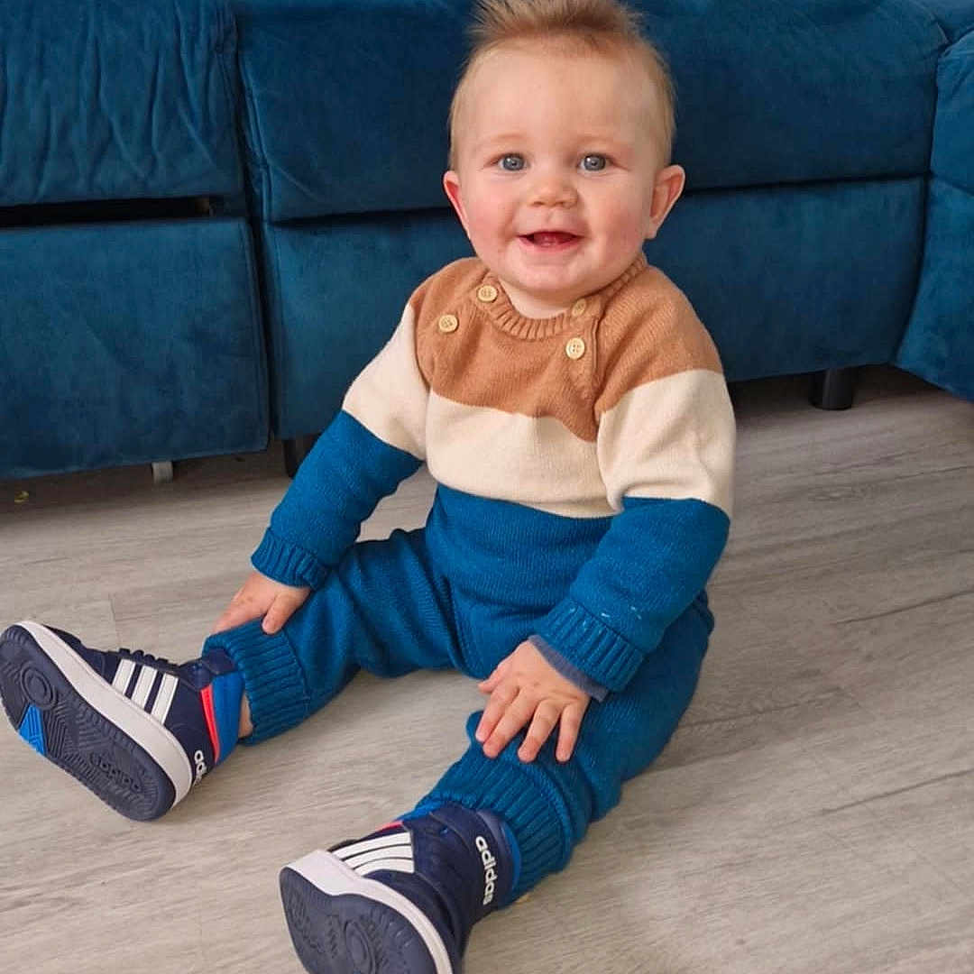 Soënn a rejoint le concours — aidez-le/la à gagner de superbes lots ! baby, blue_clothing, blue_couch, child, cozy, cute, floor, footwear, happy, home, indoor, pants, person, pillow, sitting, smiling, sneakers, spiky_hair, sweater, wooden_floor