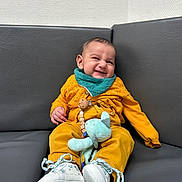 Jibrael a rejoint le concours — aidez-le/la à gagner de superbes lots ! baby, child, smiling, mustard_yellow, teal, bib, plush_toy, sneakers, couch, indoor, happy, cute, infant, clothing, seated, toy, footwear, portrait, expression, relaxed