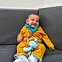 baby, child, smiling, mustard_yellow, teal, bib, plush_toy, sneakers, couch, indoor, happy, cute, infant, clothing, seated, toy, footwear, portrait, expression, relaxed