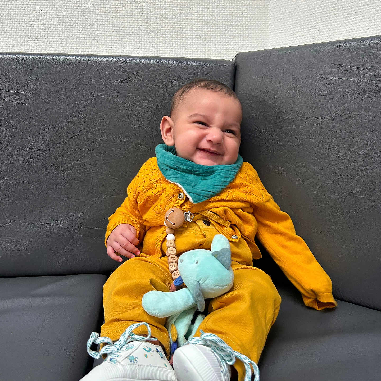 Jibrael a rejoint le concours — aidez-le/la à gagner de superbes lots ! baby, bib, child, clothing, couch, cute, expression, footwear, happy, indoor, infant, mustard_yellow, plush_toy, portrait, relaxed, seated, smiling, sneakers, teal, toy