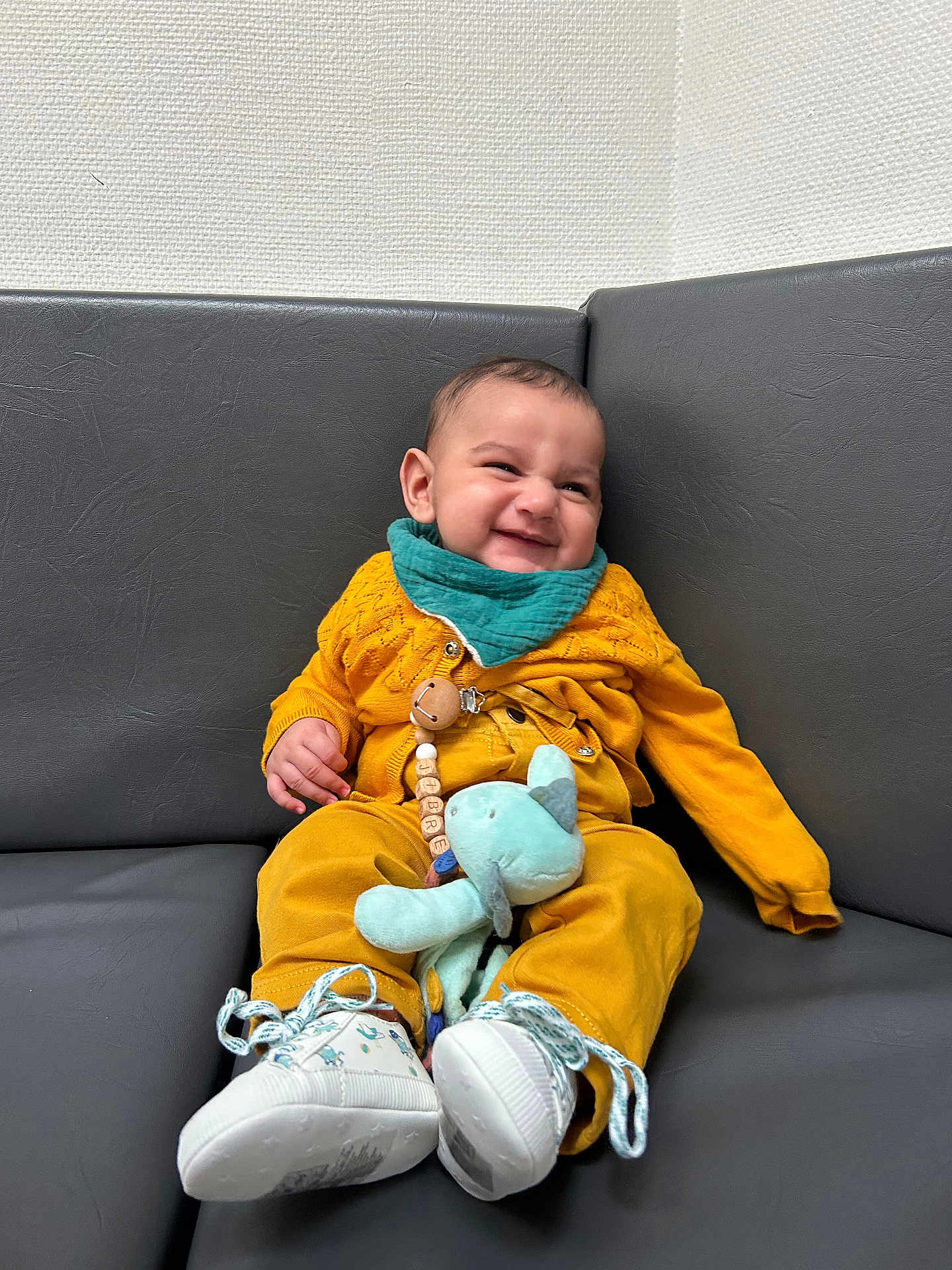 Jibrael a rejoint le concours — aidez-le/la à gagner de superbes lots ! baby, child, smiling, mustard_yellow, teal, bib, plush_toy, sneakers, couch, indoor, happy, cute, infant, clothing, seated, toy, footwear, portrait, expression, relaxed