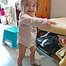 toddler, child, smiling, indoors, pigtails, white_shirt, socks, standing, table, high_chair, step_stool, wooden_floor, cabinet, toy, hand, furniture, room, happy, curly_hair, person