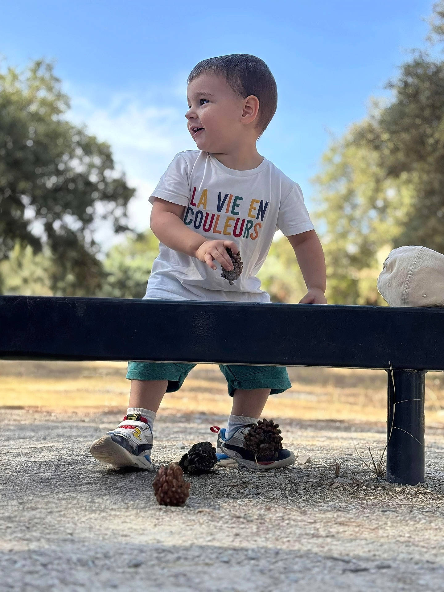 Paolo participe au concours pour gagner de l'argent avec cette photo : bench, blue_sky, child, daytime, grass, ground, happy, hat, nature, outdoor, person, pine_cone, play, shorts, smiling, sneakers, sunlight, toddler, trees, tshirt