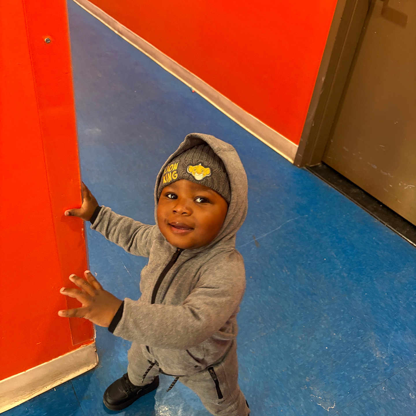 Ansyto joined the competition — help win amazing prizes! toddler, child, hoodie, beanie, lion_king, grey_clothing, orange_wall, blue_floor, indoor, door, curious, smiling, standing, black_shoes, person, young_child, flooring, wall, casual_wear, playful