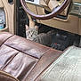 cat, tabby, vehicle_interior, steering_wheel, car_seat, leather_seat, dashboard, floor_mat, pedal, water_bottle, pet, curious_cat, whiskers, green_eyes, close_up, upholstery, front_seat, passenger_area, indoor, automobile