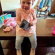 Rivhur is registered to the contest to win money with this photo: toddler, child, pink_headband, pink_top, jeans, boots, wooden_table, phone_case, rocking_chair, blanket, diapers_box, carpet, indoor, curious_expression, child_sitting, home, furniture, young_child, casual_clothing, playful