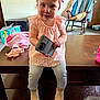 toddler, child, pink_headband, pink_top, jeans, boots, wooden_table, phone_case, rocking_chair, blanket, diapers_box, carpet, indoor, curious_expression, child_sitting, home, furniture, young_child, casual_clothing, playful