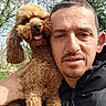 man, dog, outdoor, portrait, smiling, curly_hair, brown_dog, nature, trees, path, sunlight, happy, close_up, pet, animal, person, face, canine, fur, jackets