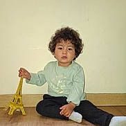 Anouar a rejoint le concours — aidez-le/la à gagner de superbes lots ! black_pants, casual_clothing, child, curly_hair, eiffel_tower, floor, golden, green_shirt, indoor, model, neutral_expression, patterned_rug, rug, sitting, socks, toddler, toy, wall, window, wooden_floor