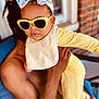 baby, child, sunglasses, headband, bow, yellow_outfit, bib, bee_design, adult_hand, portrait, outdoor, window, brick_wall, fashion, cute, person, holding, infant, cool, style
