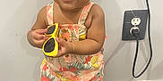Ziya is registered to the contest to win money with this photo: toddler, child, baby, standing, yellow_sunglasses, floral_romper, white_sandals, bow, hair_bow, carpet, indoor, wall, electrical_outlet, cord, smiling, cute, person, portrait, holding, feet