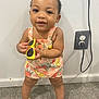toddler, child, baby, standing, yellow_sunglasses, floral_romper, white_sandals, bow, hair_bow, carpet, indoor, wall, electrical_outlet, cord, smiling, cute, person, portrait, holding, feet