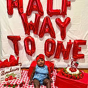 Kaira is registered to the contest to win money with this photo: baby, child, balloons, strawberries, cake, headband, denim_overalls, red, checkered_blanket, celebration, milestone, smiling, indoor, crate, food, fruit, decorations, birthday, sitting, happy