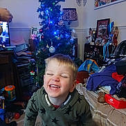 Thomas-james joined the competition — help win amazing prizes! cheerful, child, christmas_tree, clothing, decorations, face, festive, furniture, green_hoodie, holiday, home, indoor, lamp, living_room, person, pile_of_clothes, playful, smiling, toddler, toy