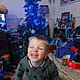 cheerful, child, christmas_tree, clothing, decorations, face, festive, furniture, green_hoodie, holiday, home, indoor, lamp, living_room, person, pile_of_clothes, playful, smiling, toddler, toy